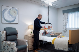 Carer is next to the client, who is sitting on a bed in a healthcare sling. The sling is attached to a ceiling track hoist.