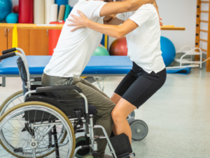 Somenone being helped up from a wheelchair. The wheelchair user is placing their arms around the carers neck. The carer has their arms either side of the users torso.