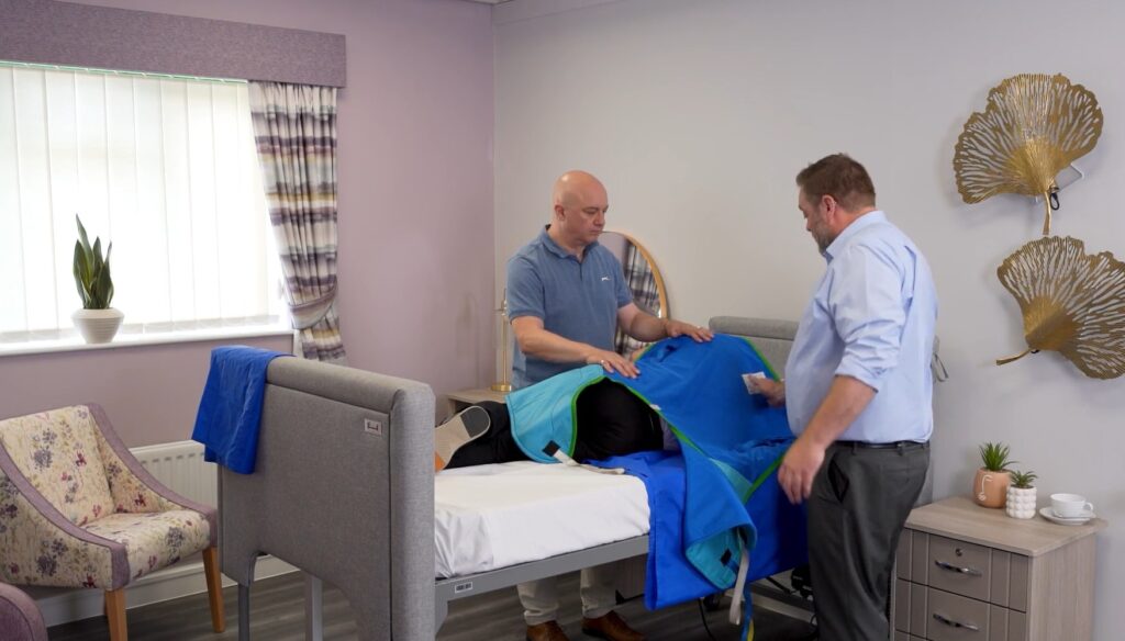 Two caregivers are applying a Prism Medical UK sling to a man lying on his side in bed. This is a demonstration from a moving and handling training webinar.